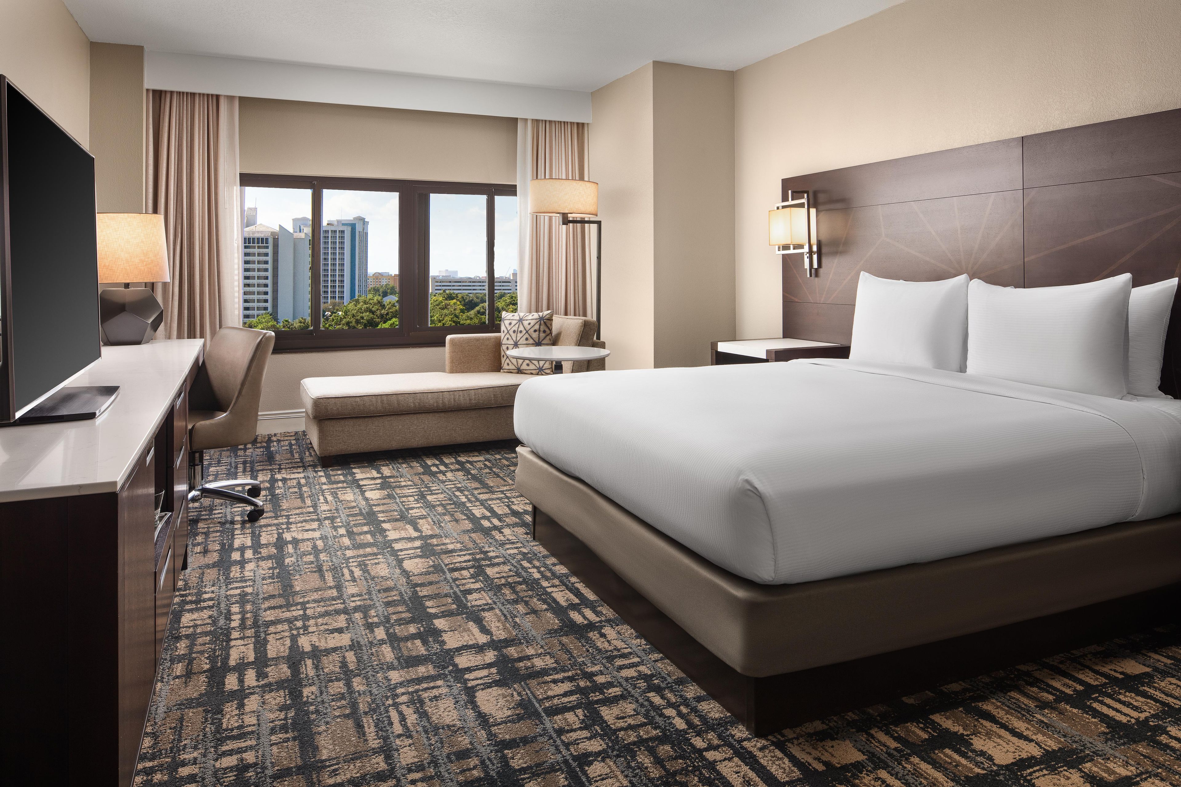 A hotel room with a large bed, a desk, a chair, a small table, and a view of the cityscape.
