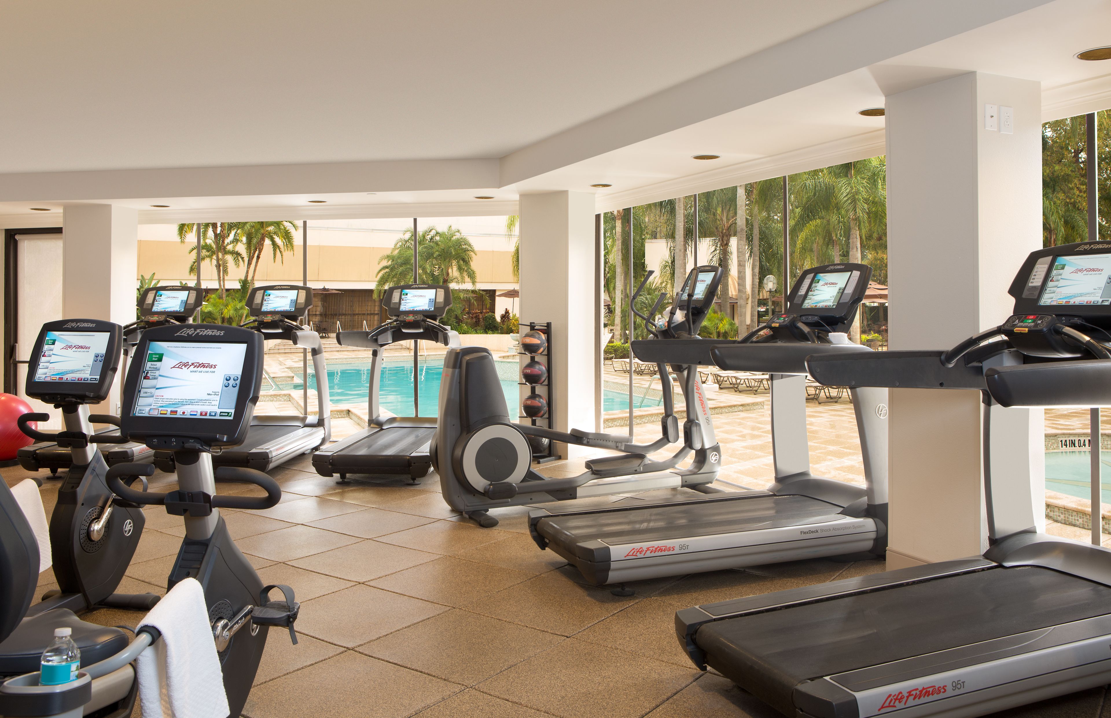 A gym with treadmills, stationary bikes, and a pool area visible through large windows.
