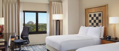 Habitación de hotel con dos camas, un escritorio con silla, lámpara, reloj y ventana con vistas a edificios y árboles.