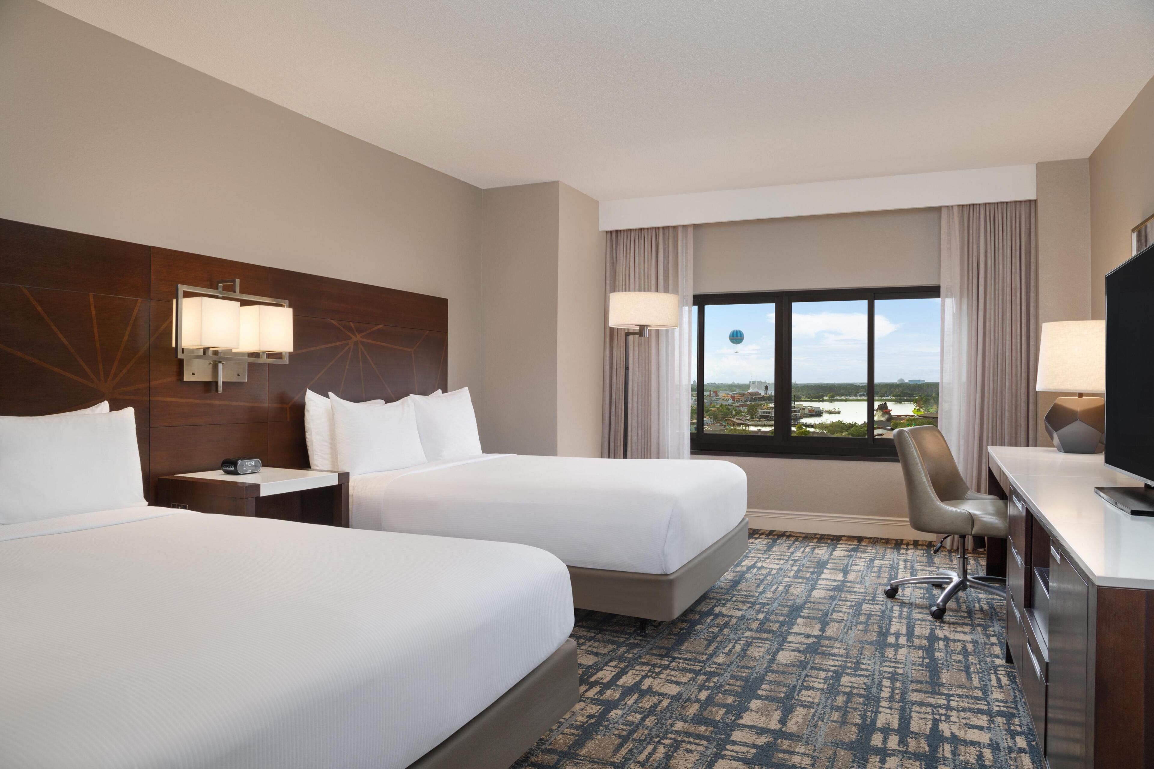 A hotel room with two beds, a desk, a chair, a TV, and a window with a city view.