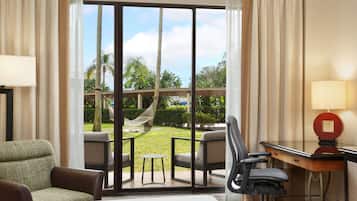 Habitación de hotel con cama, escritorio con lámpara, una silla y vista a un jardín con hamaca.