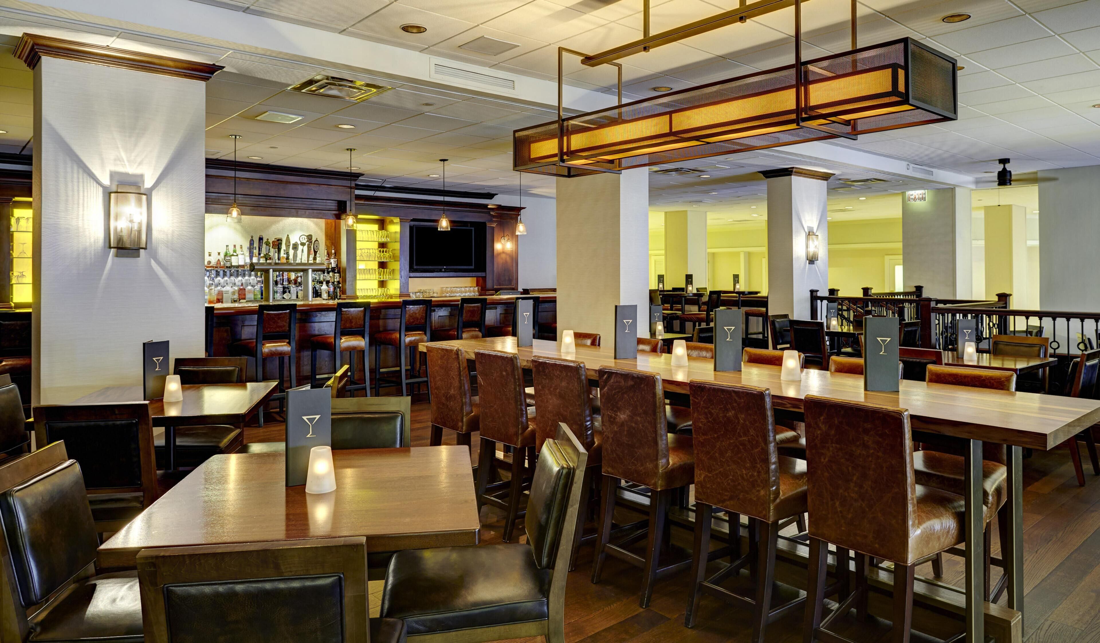 A restaurant with multiple dining tables and chairs, a bar area with bottles and glasses, and a television screen.