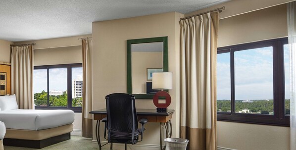 A hotel room with a bed, desk, chair, lamp, and large windows offering a city view.