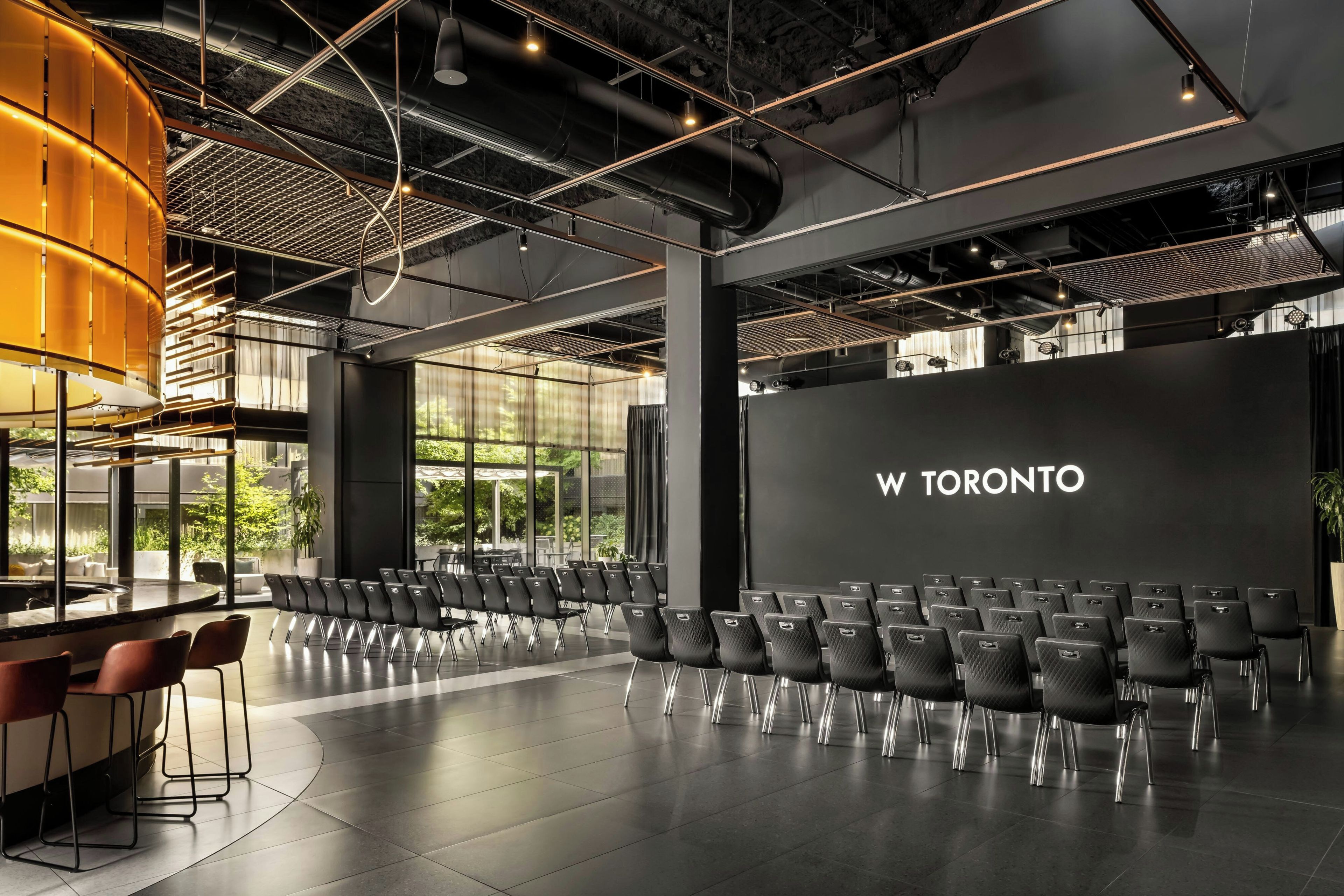 Una sala de conferencias moderna con filas de sillas, una pantalla grande que muestra 'W TORONTO' y una zona de bar con taburetes.