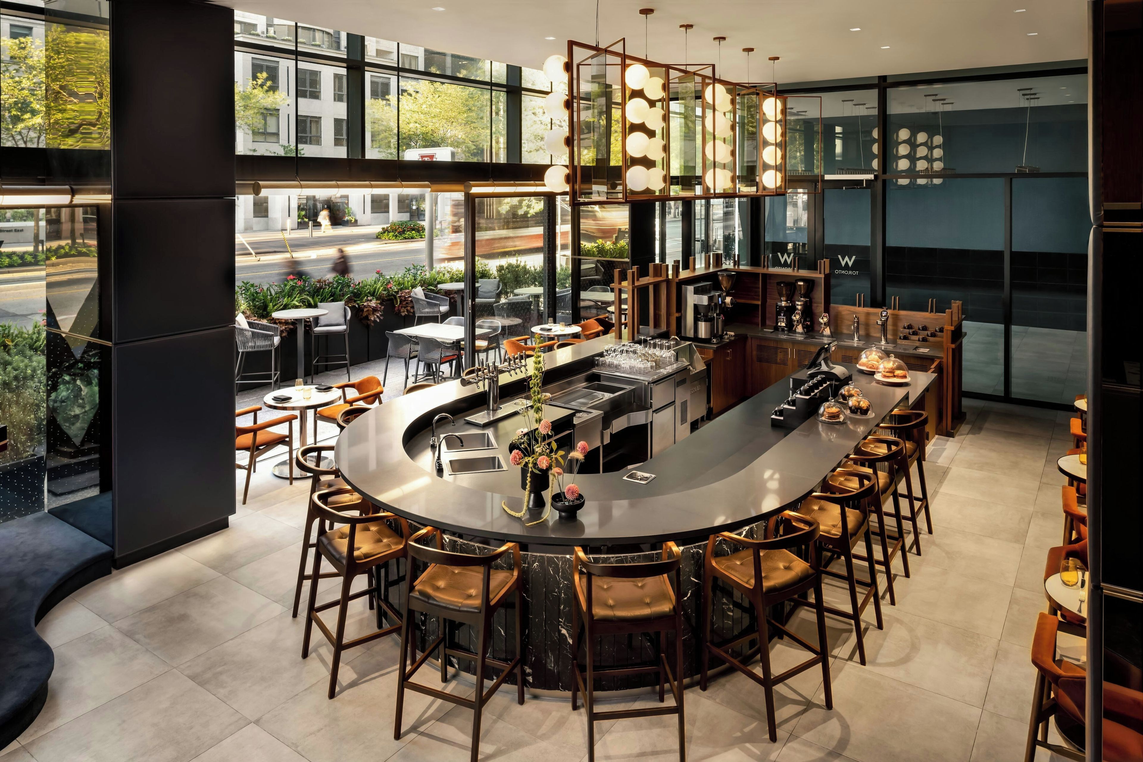 A modern bar area with a curved counter, bar stools, and a view of the street through large windows.