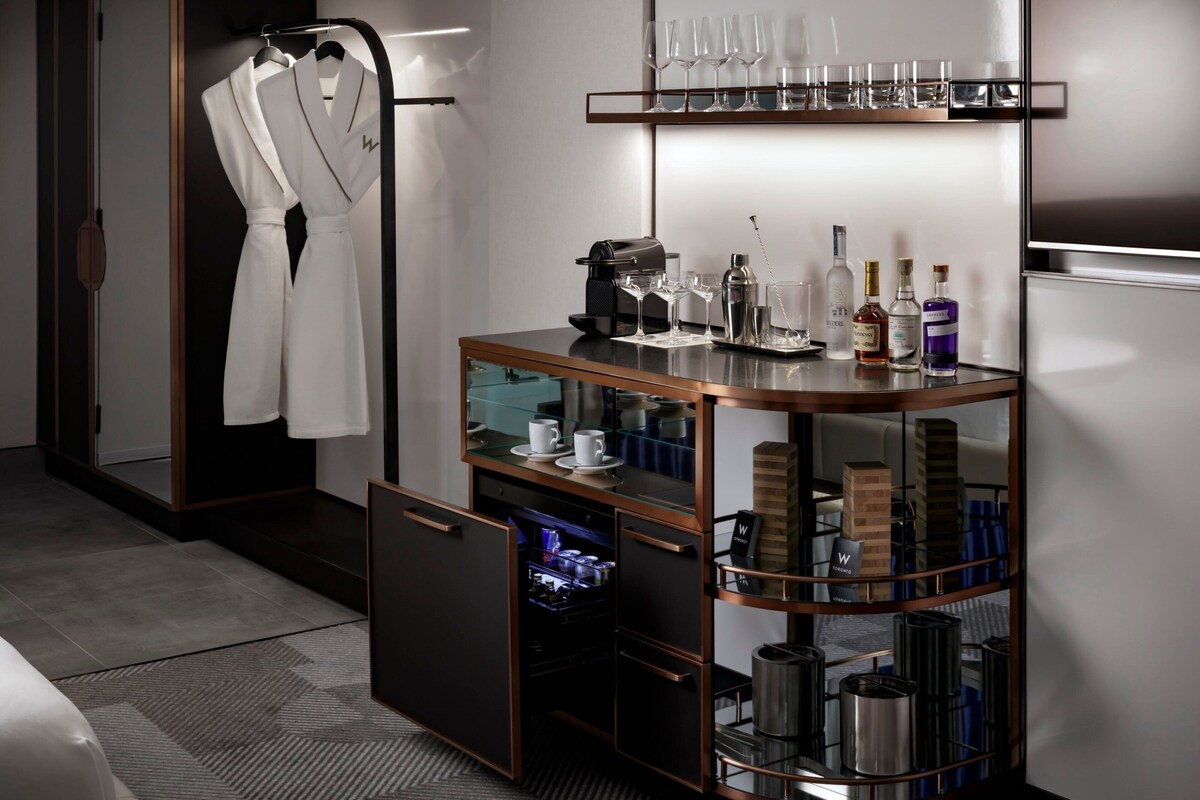 A hotel room with a minibar, a glass display case with bottles, a coffee machine, and a hanging robe.