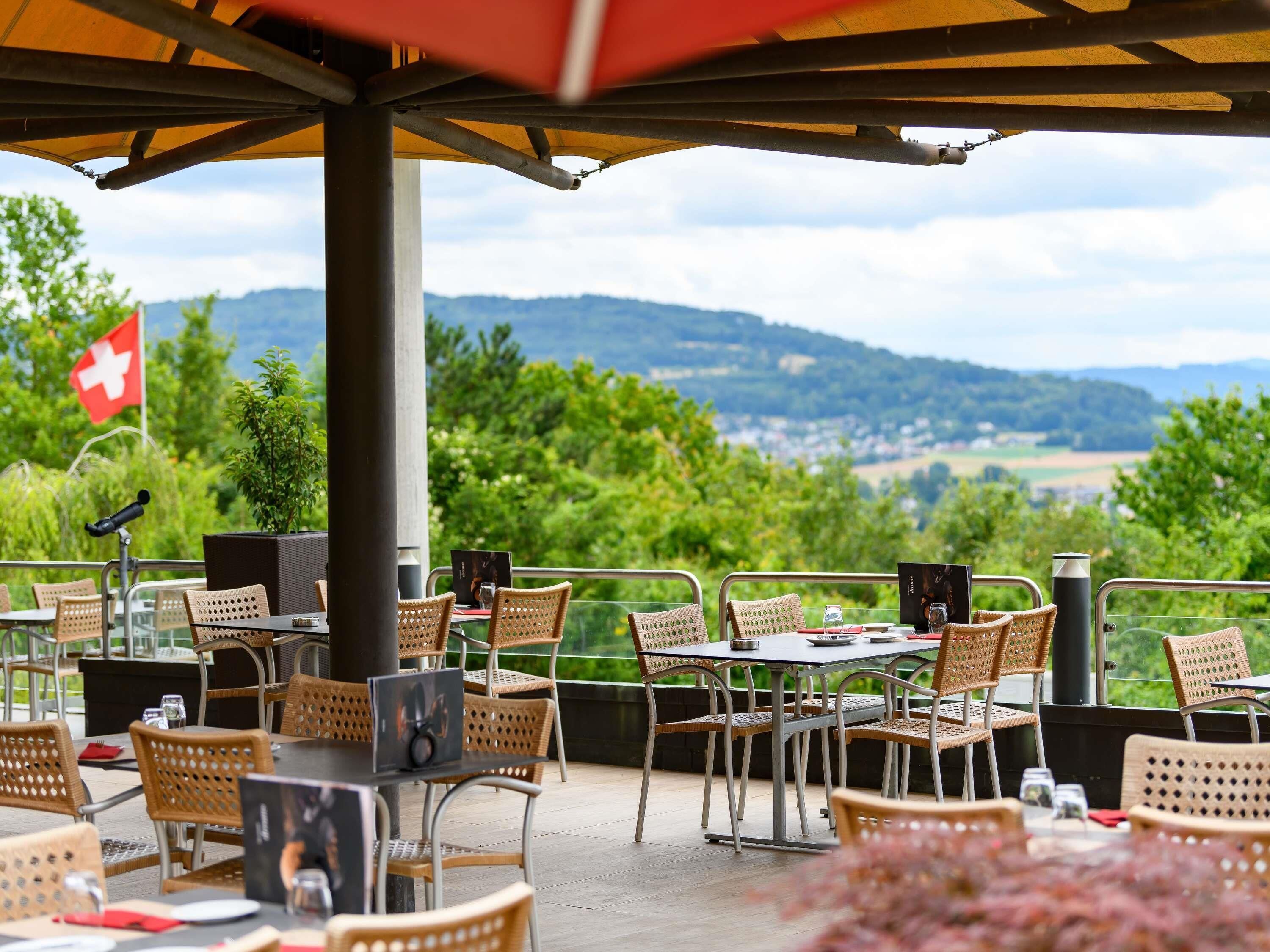 An outdoor dining area with tables and wicker chairs, a Swiss flag, and a view of the countryside.