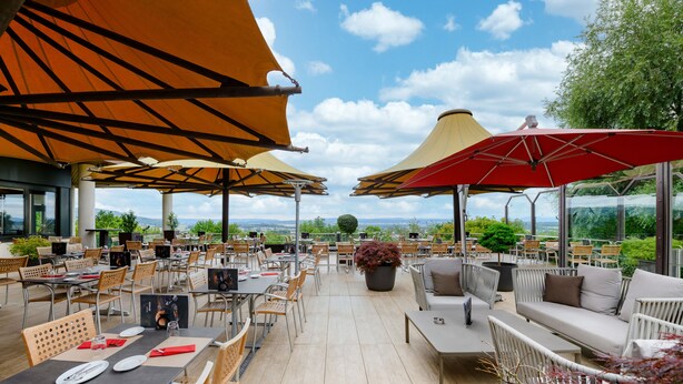 Un espacio de comedor al aire libre con mesas y sillas, grandes sombrillas y vistas del paisaje.