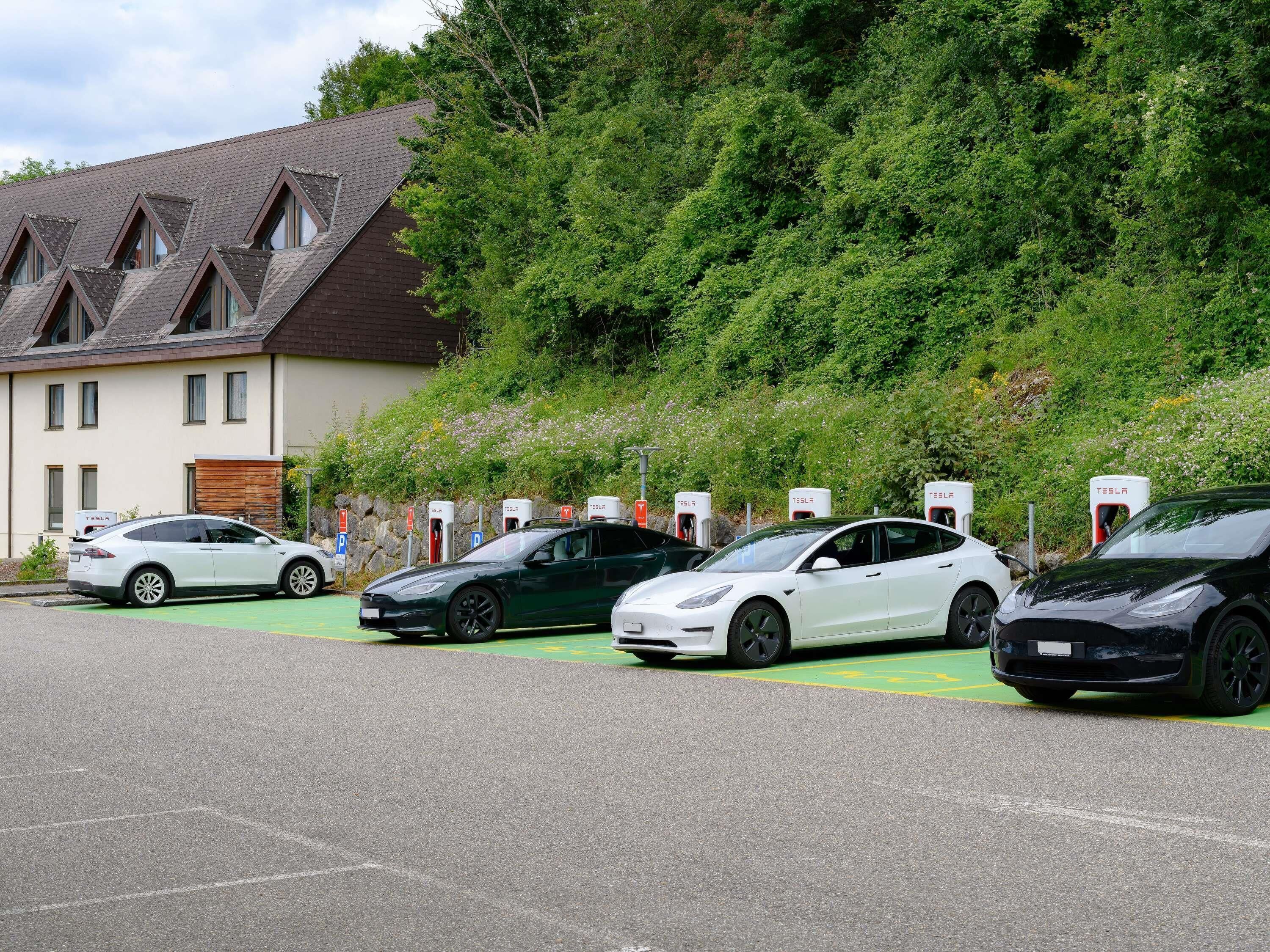 Uma área de estacionamento com estações de carregamento Tesla, um edifício com telhado inclinado e um morro coberto por vegetação densa.