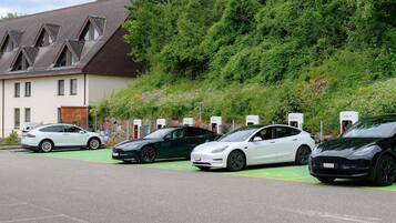 Uma área de estacionamento com estações de carregamento Tesla, um edifício com telhado inclinado e um morro coberto por vegetação densa.