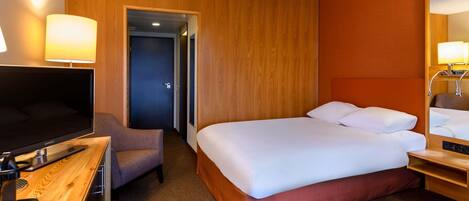A hotel room with a bed, a desk with a TV, a chair, a bedside table, and a mirror.