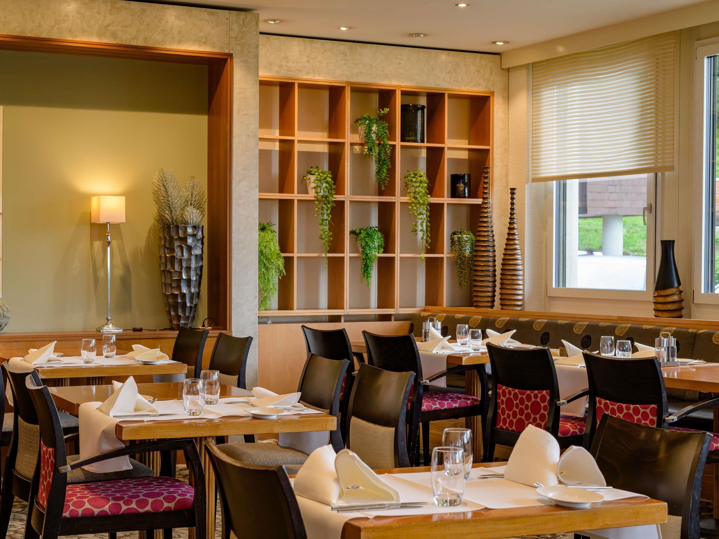 A restaurant with wooden tables and chairs, a large plant display, and a window with blinds.
