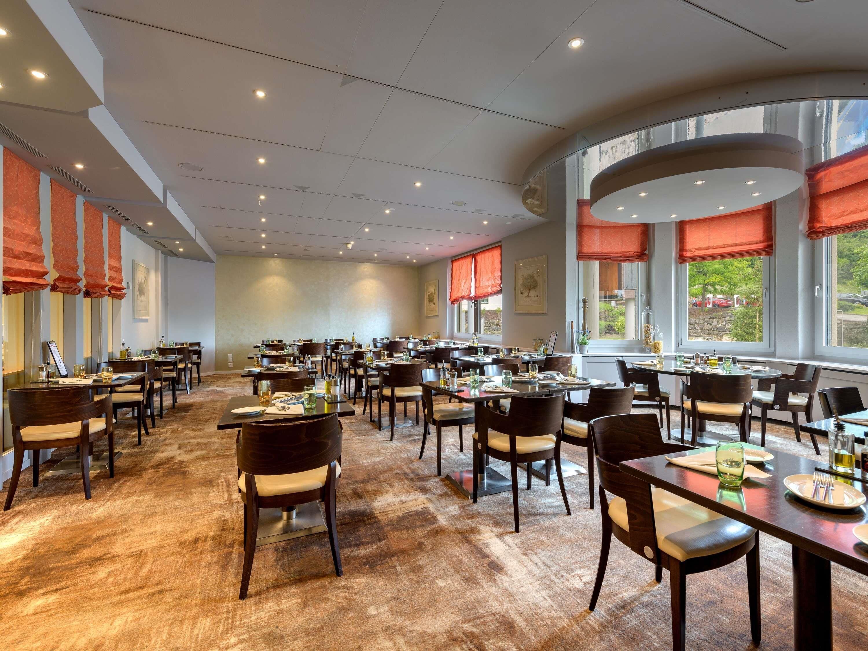 A spacious dining area with multiple tables and chairs, large windows with red valances, and a high ceiling with recessed lighting.