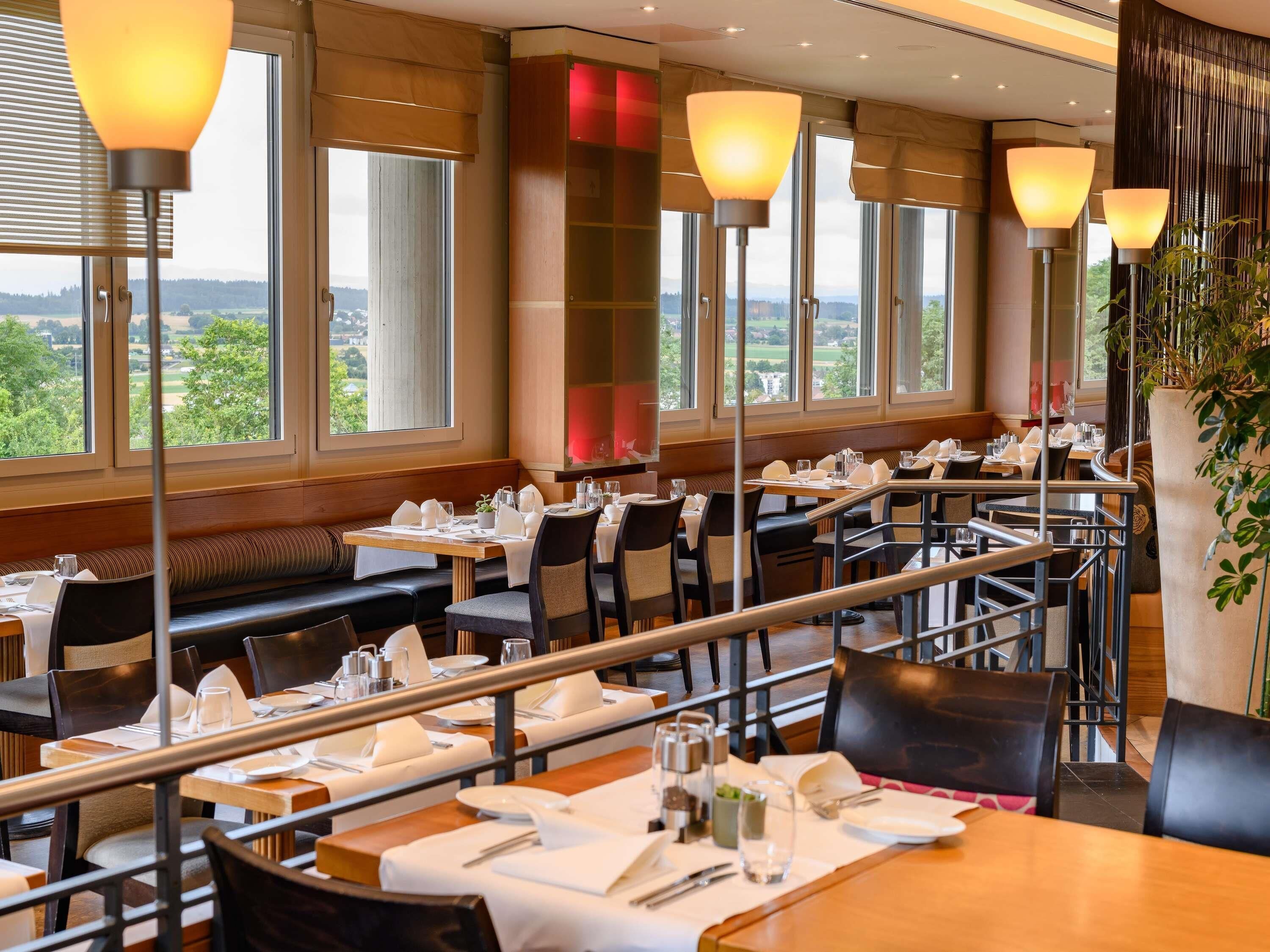 Un restaurant doté de nombreuses tables et de banquettes, de grandes fenêtres donnant sur l’extérieur et d’un éclairage moderne.