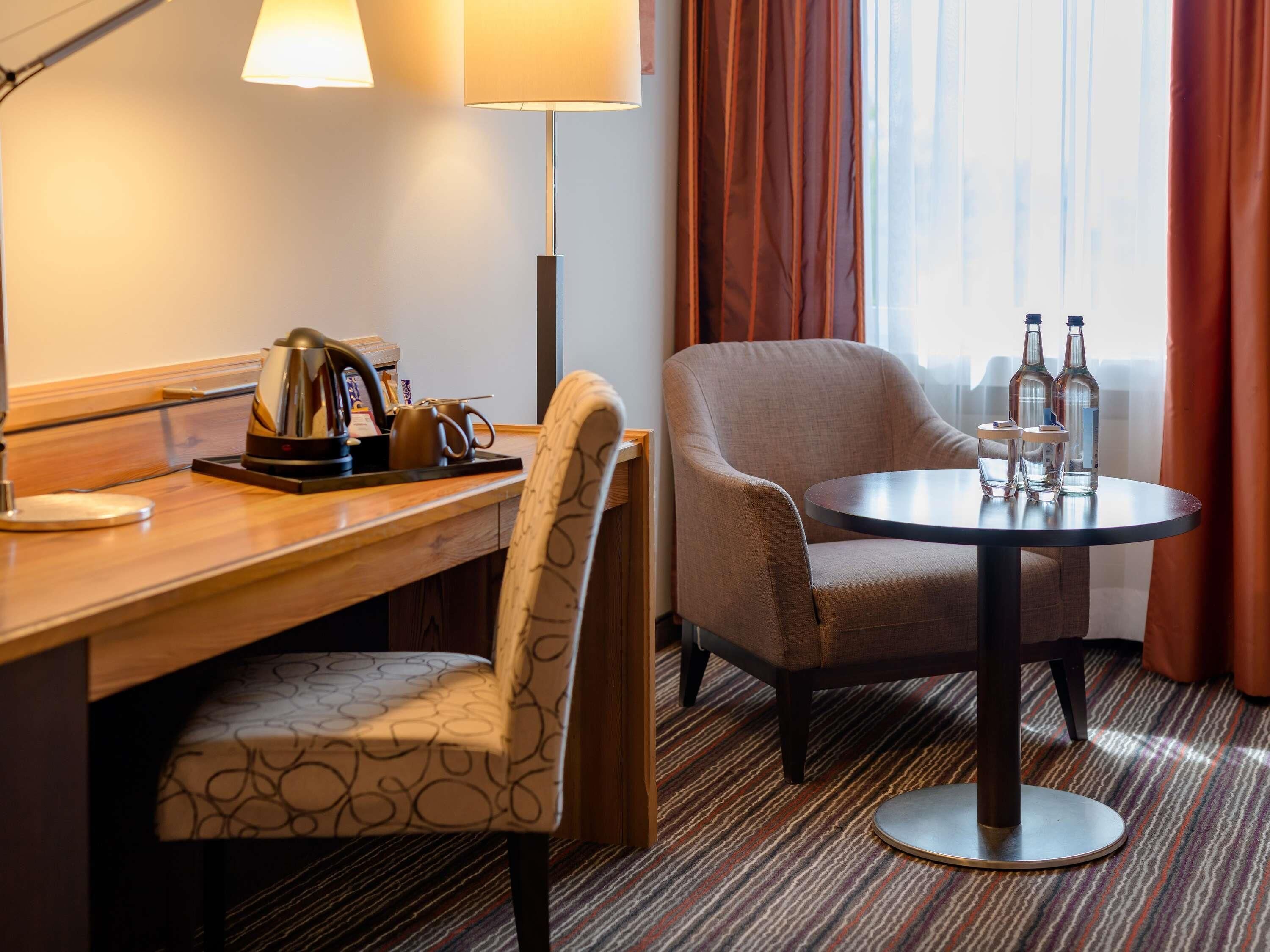 A hotel room with a desk, a chair, a round table with bottles, a lamp, and a window with curtains.