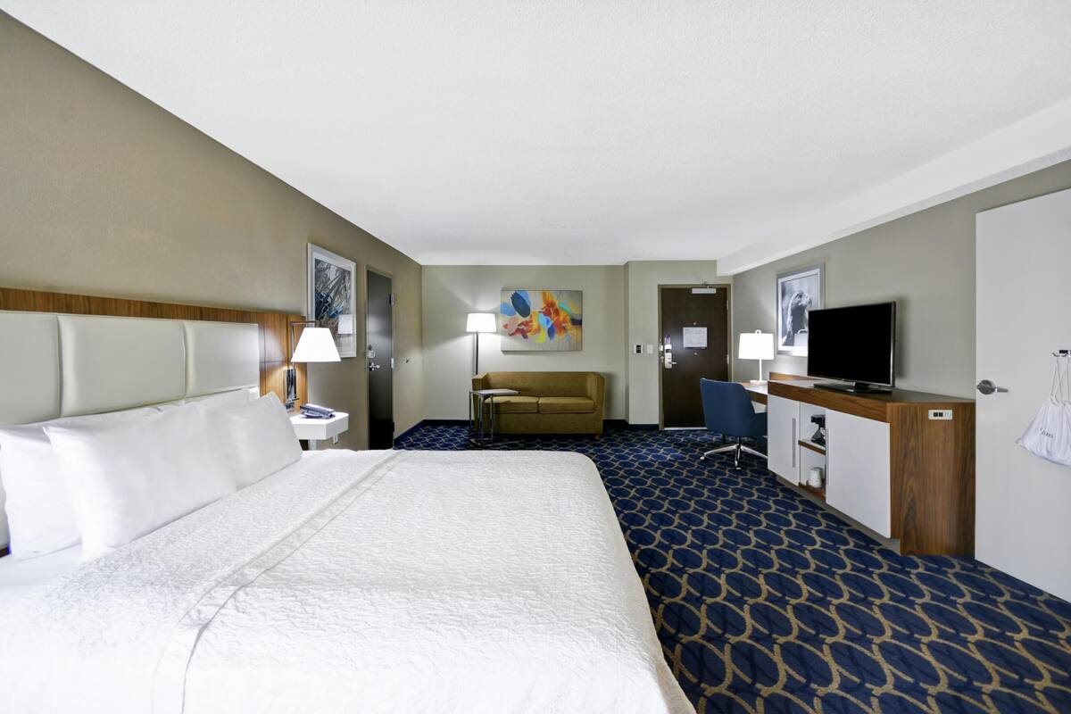 A hotel room with a large bed, a television, a desk, a chair, a sofa, and a painting on the wall.