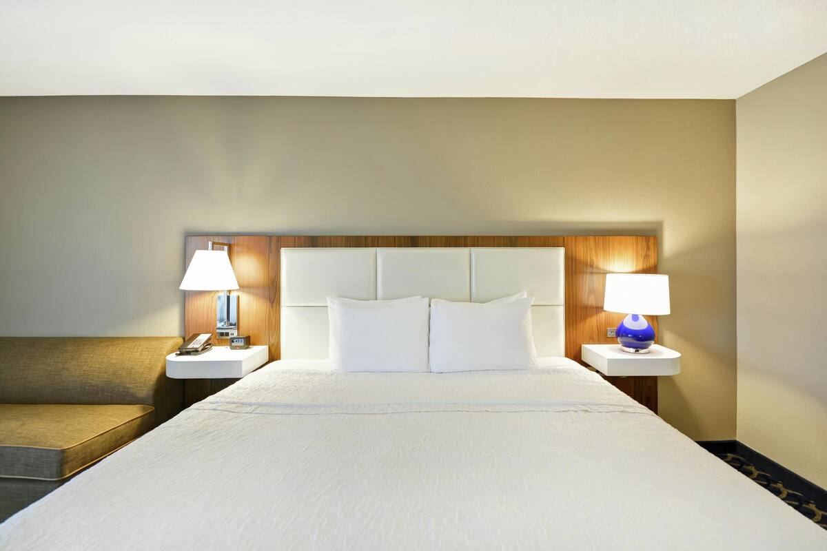A hotel room with a large bed, two bedside tables, a nightstand, a lamp, and a sofa.