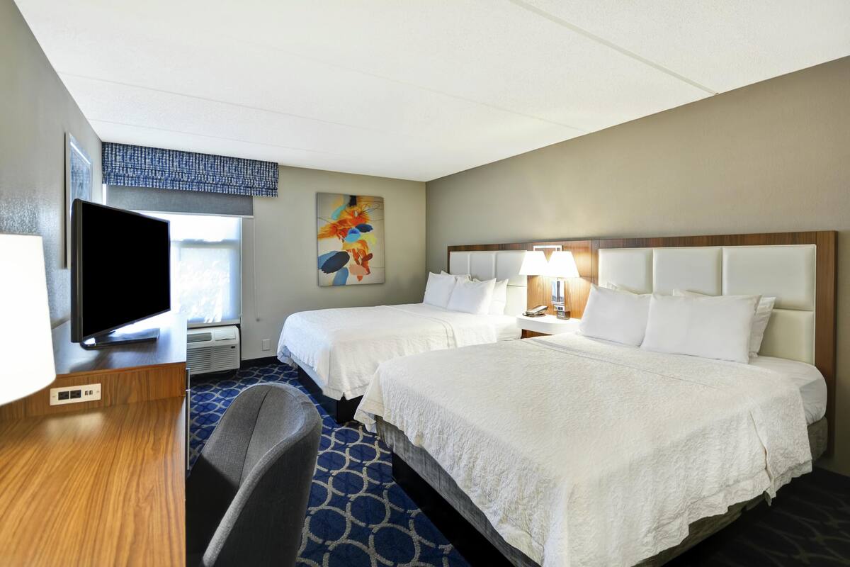 A hotel room with two beds, a television, a painting, and a window with blinds.