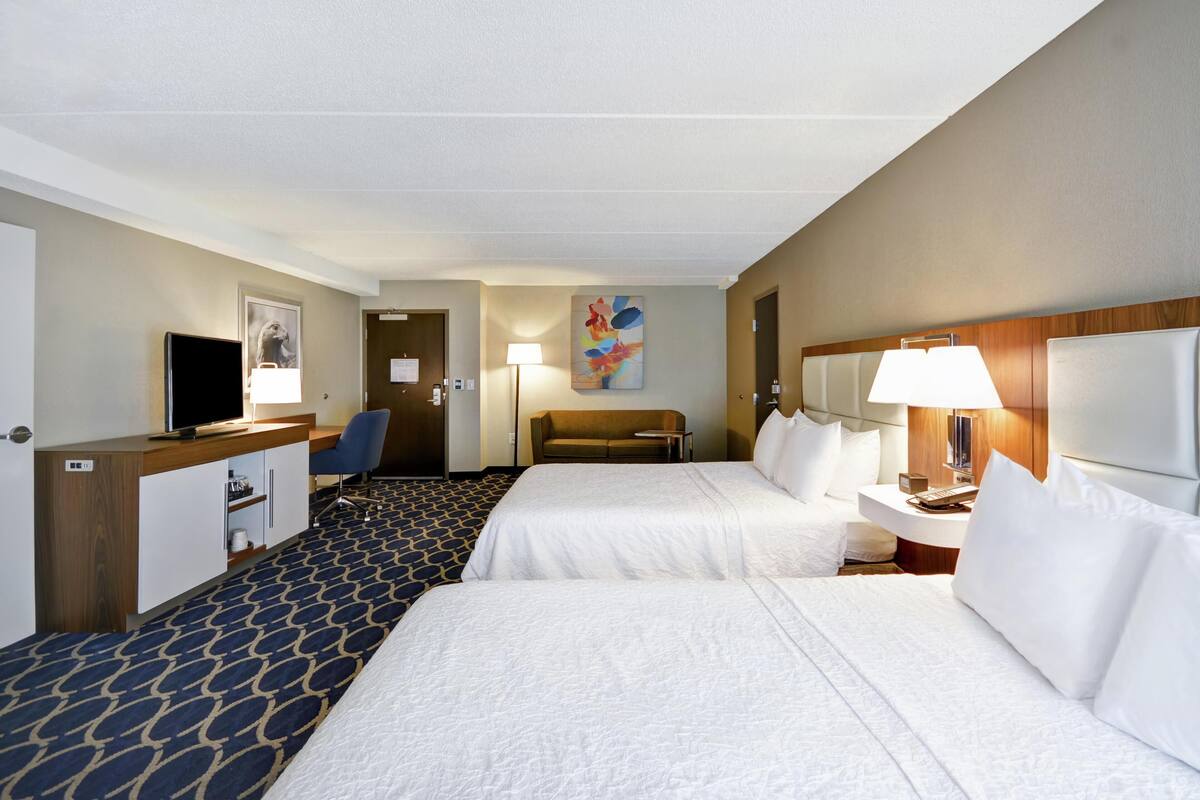 A hotel room with two beds, a desk, a TV, a sofa, and a painting on the wall.