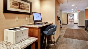 Zona de recepción de hotel con un ordenador de escritorio, una impresora, una fotografía enmarcadas en la pared y un pasillo que conduce a la salida.