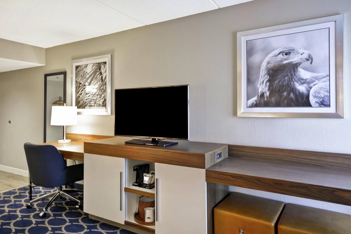 A hotel room with a desk, a television, a coffee machine, a chair, and framed artwork on the wall.