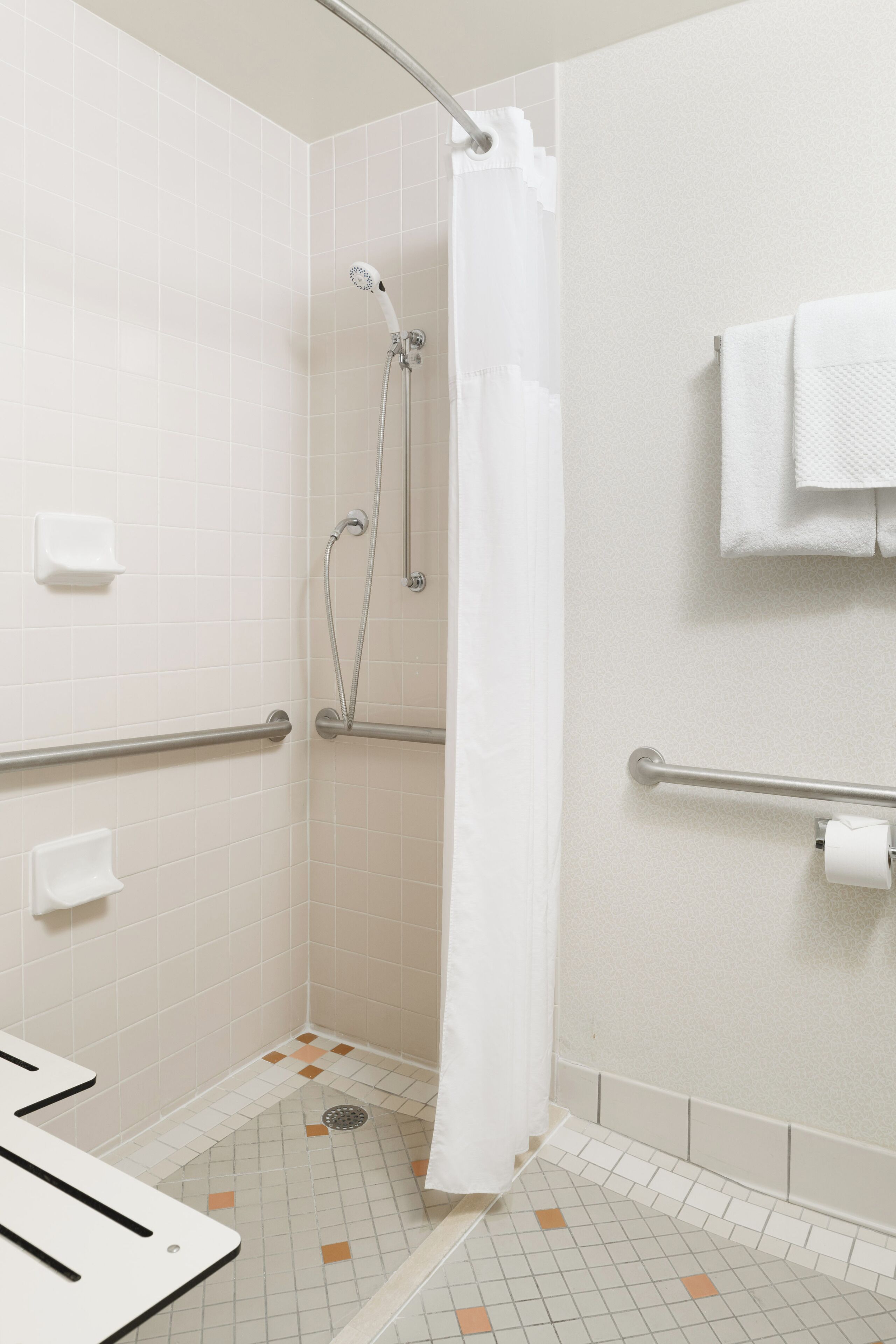 Un baño con paredes de azulejos blancos, una ducha con cortina blanca, una barra de apoyo, un porta papel higiénico y un perchero con una toalla.