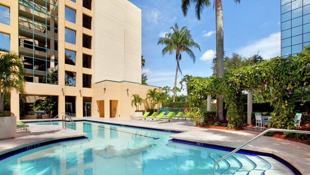 Ein Pool mit gefliestem Rand, umgeben von Liegestühlen und Palmen, mit einem modernen Gebäude im Hintergrund.
