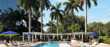 A swimming pool with lounge chairs and umbrellas, surrounded by palm trees and a modern building.