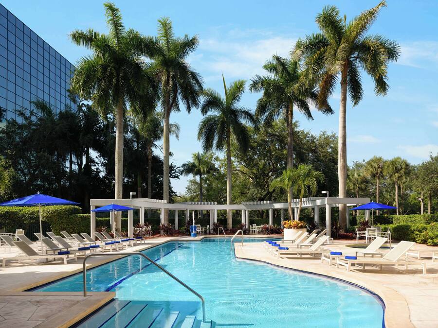 A swimming pool with lounge chairs and umbrellas, surrounded by palm trees and a modern building.