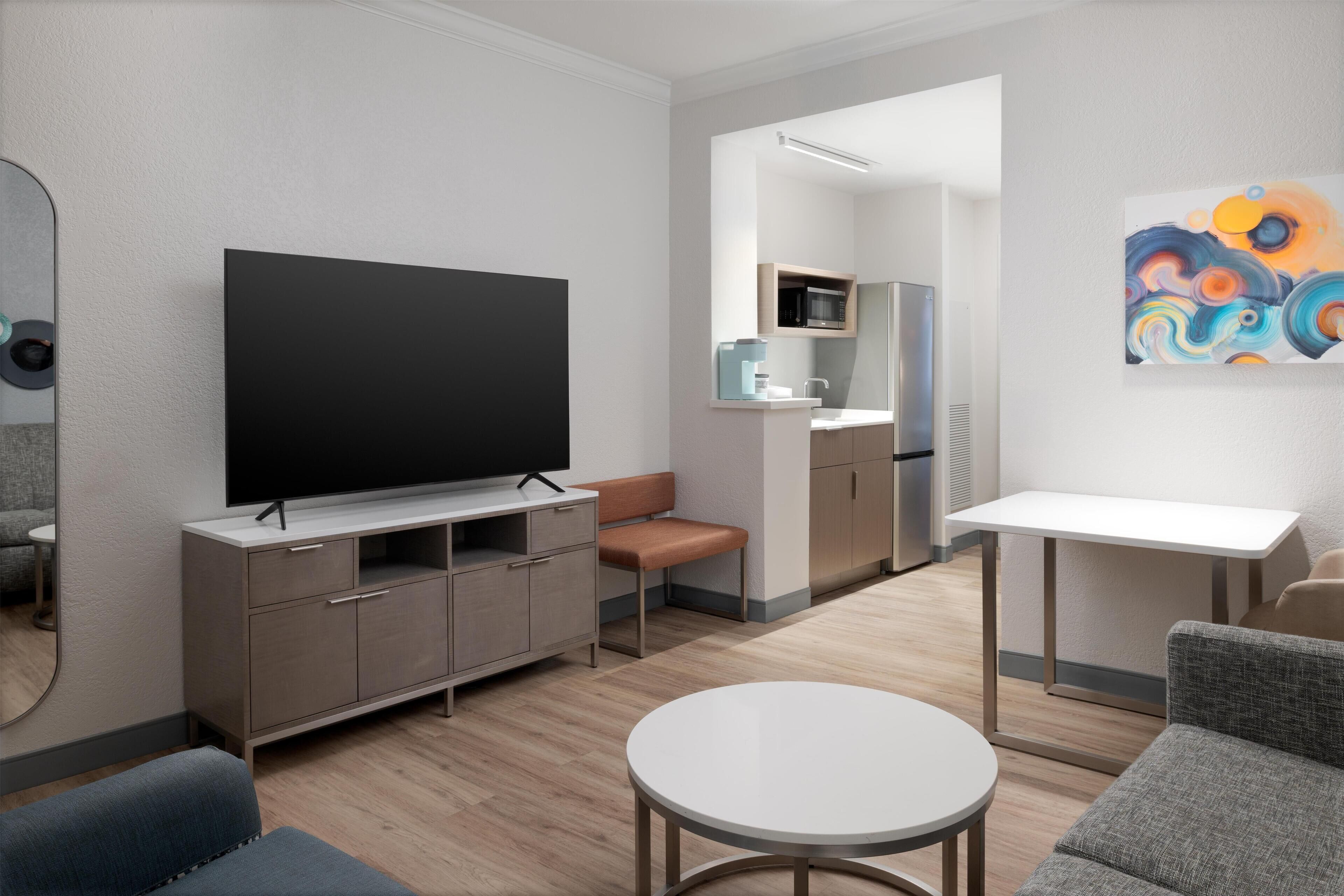 A modern living room with a flat-screen TV, a sofa, a coffee table, and a kitchen area in the background.