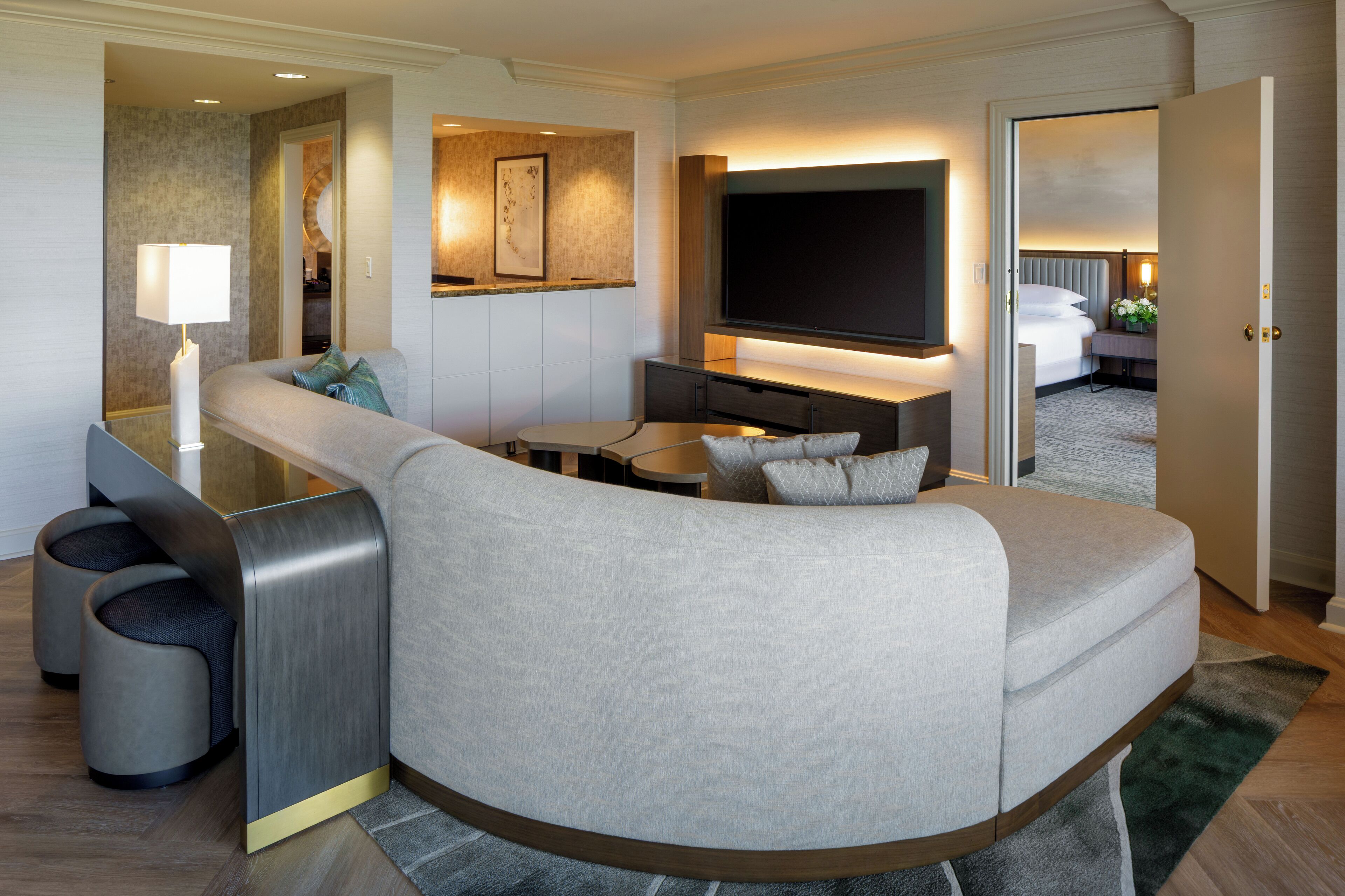 A modern hotel room with a curved sofa, a flat-screen TV, a desk, and a separate sleeping area.