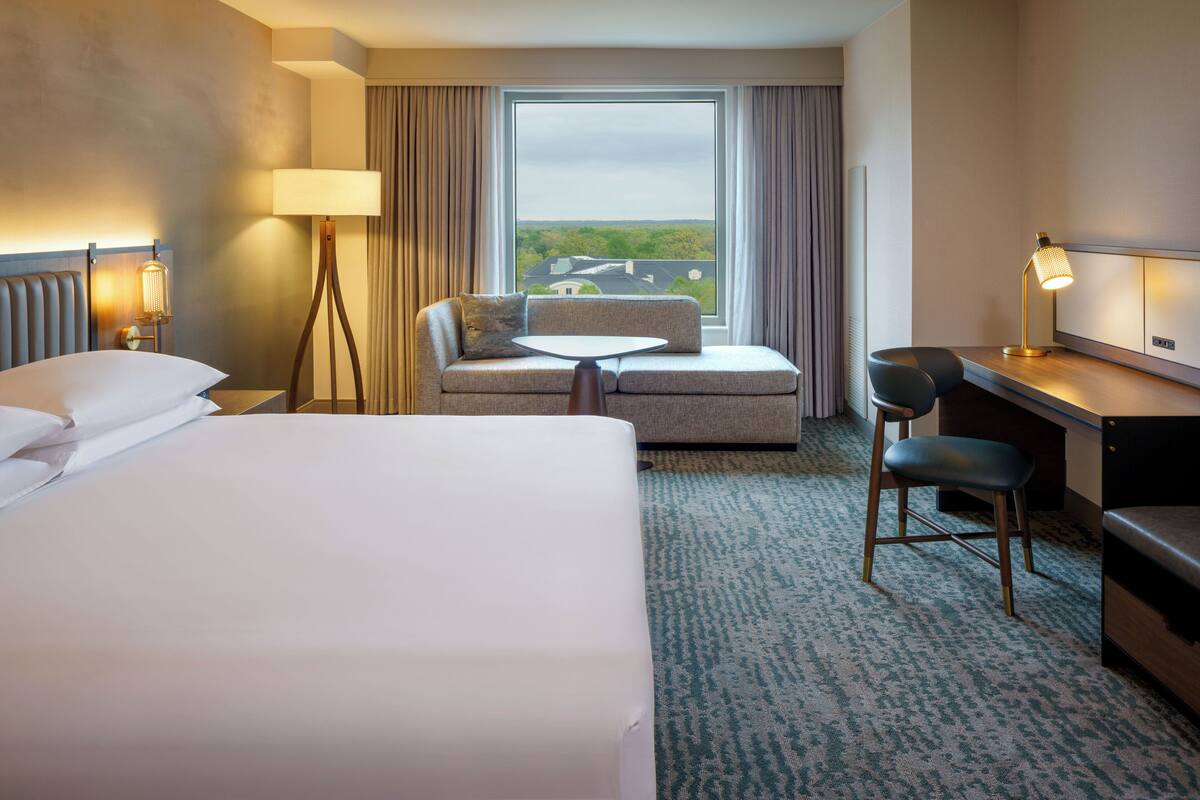 Ein Hotelzimmer mit Bett, Sofa, einem kleinen Tisch, einem Schreibtisch und Blick ins Freie.