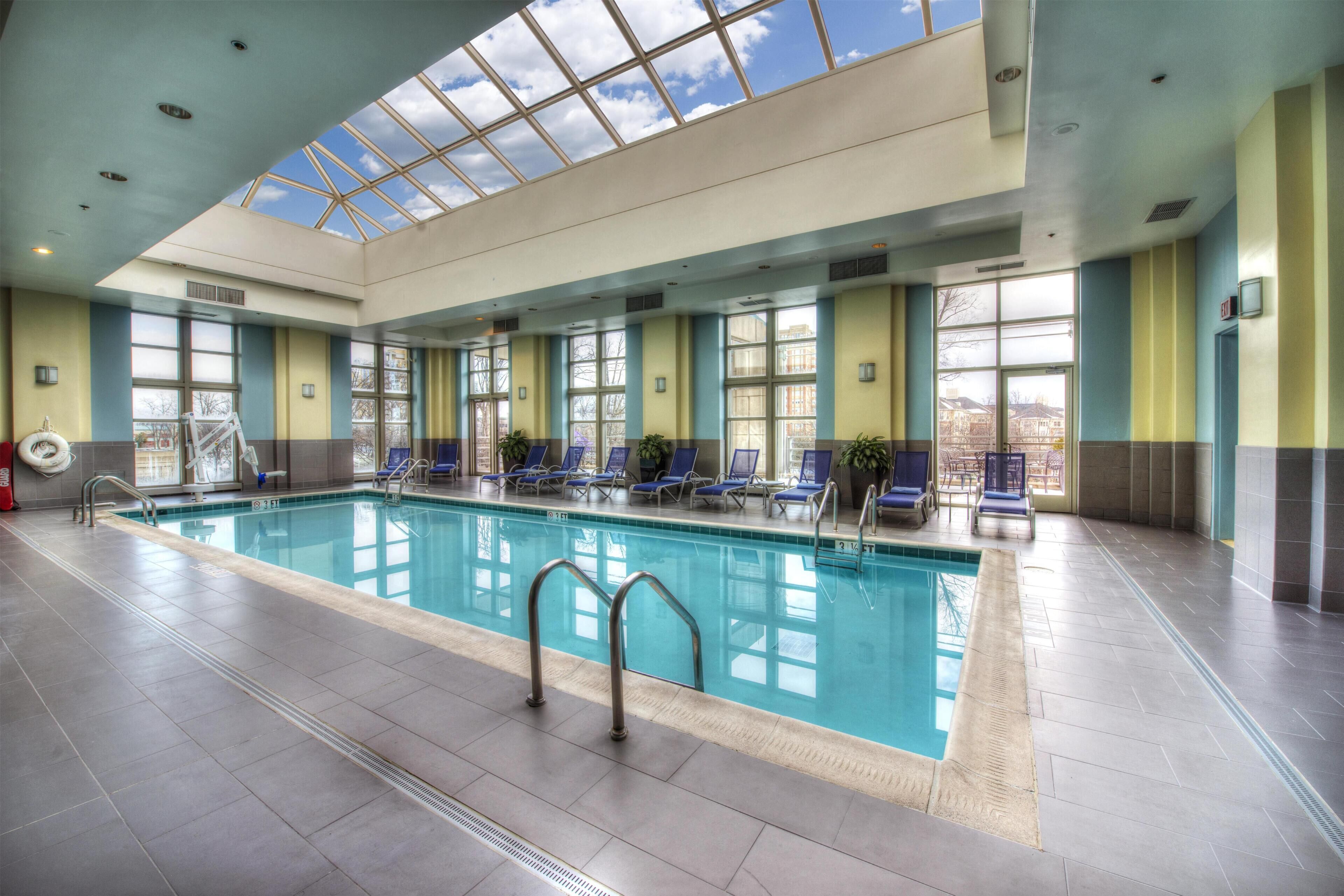 An indoor swimming pool with clear glass ceiling, blue lounge chairs, and a fire extinguisher.