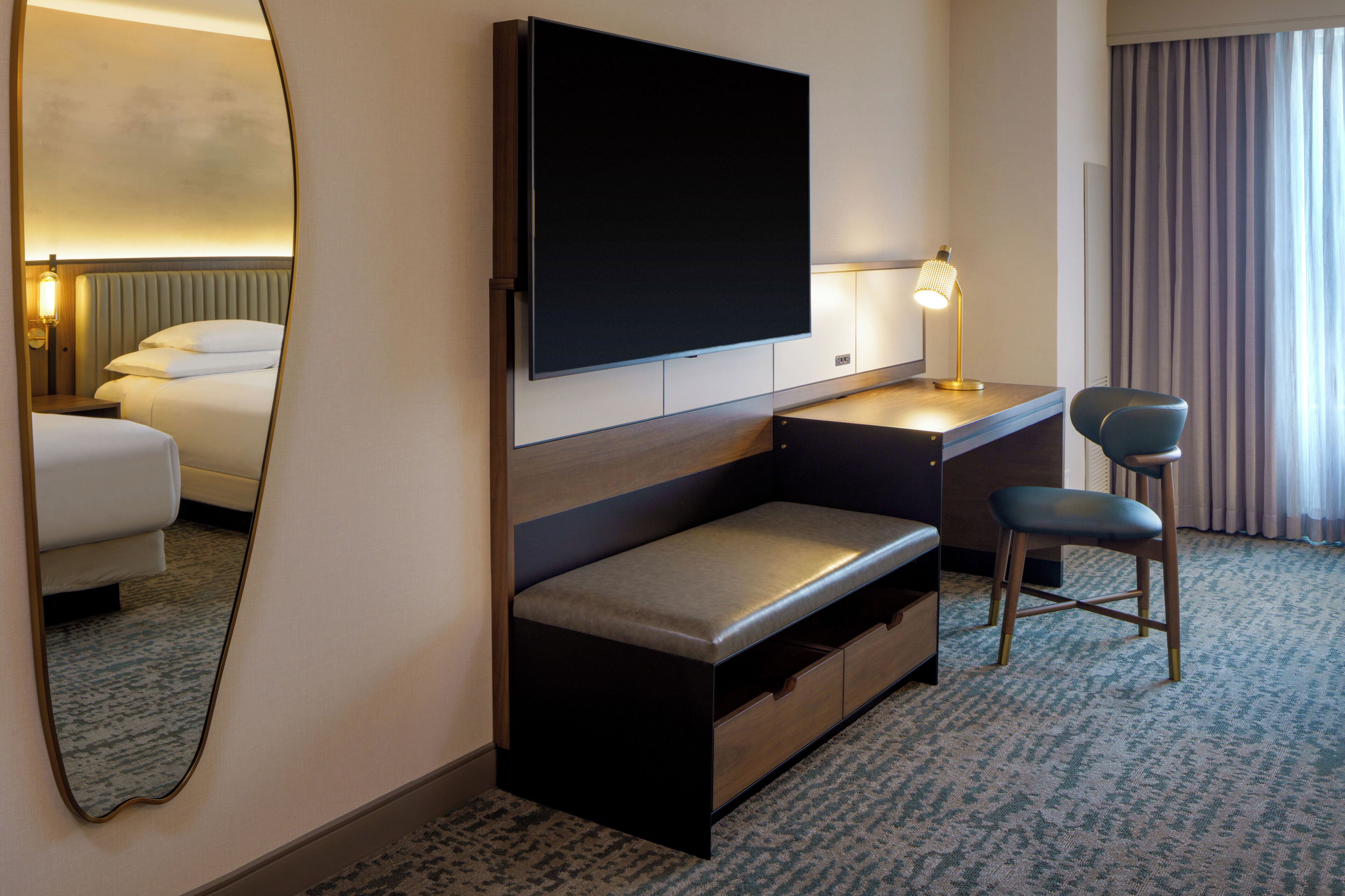 A hotel room with a bed, a desk with a chair, a flat-screen TV, a bedside lamp, and a mirror.