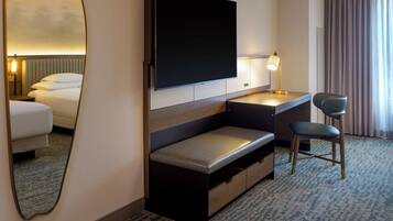 Quarto de hotel com uma cama, uma escrivaninha com cadeira, uma TV de tela plana, um abajur e um espelho.