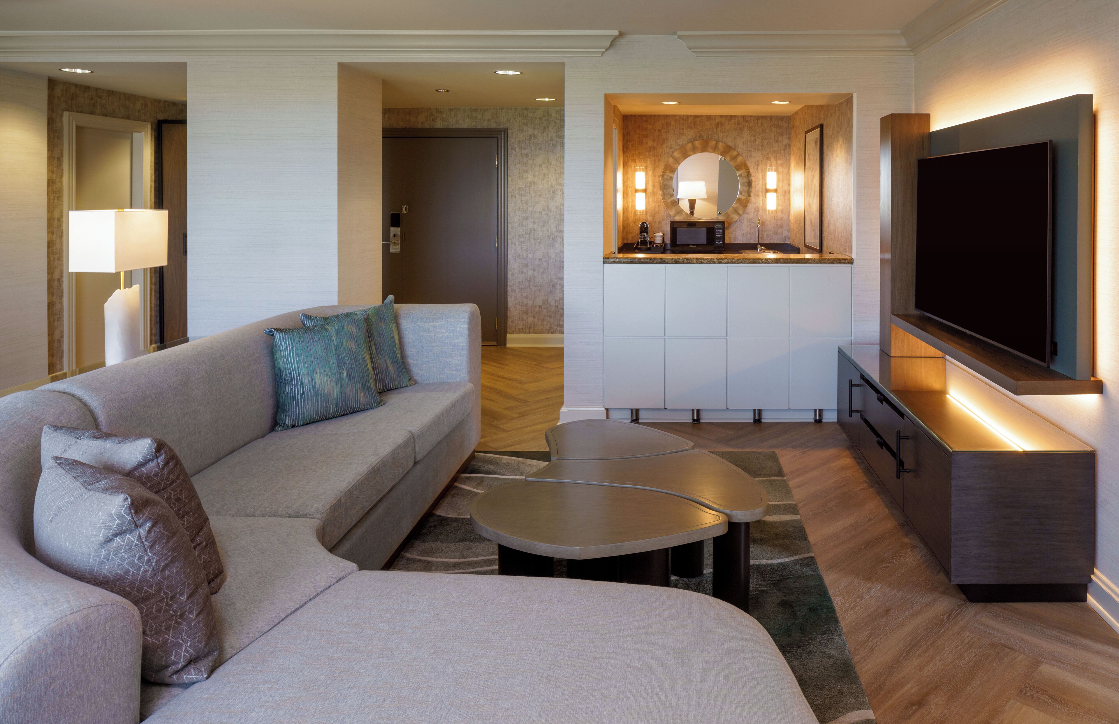 A modern hotel room with a sofa, two small tables, a flat-screen TV, a kitchen area with a microwave, and a lamp.