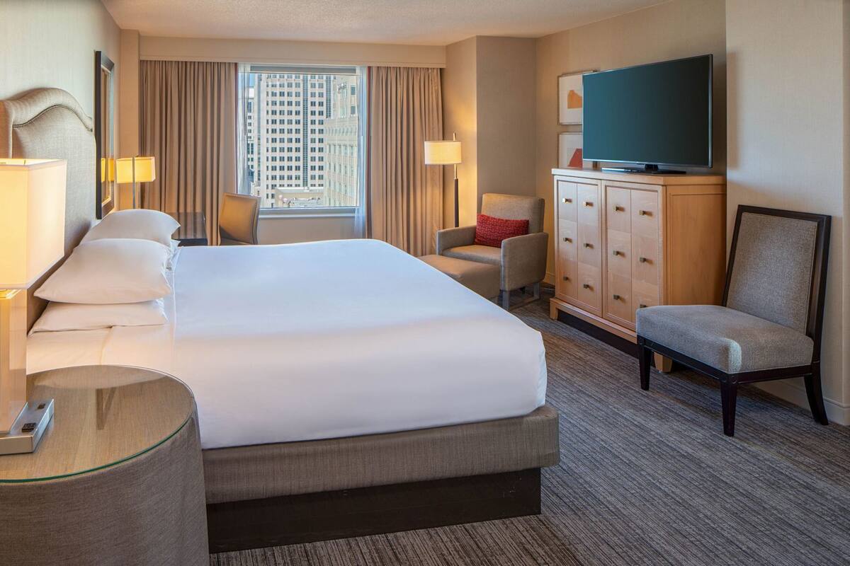 Ein Hotelzimmer mit einem großen Bett, einem Fernseher, einem Schreibtisch mit Lampe, einem Sessel und einem Kleiderschrank.