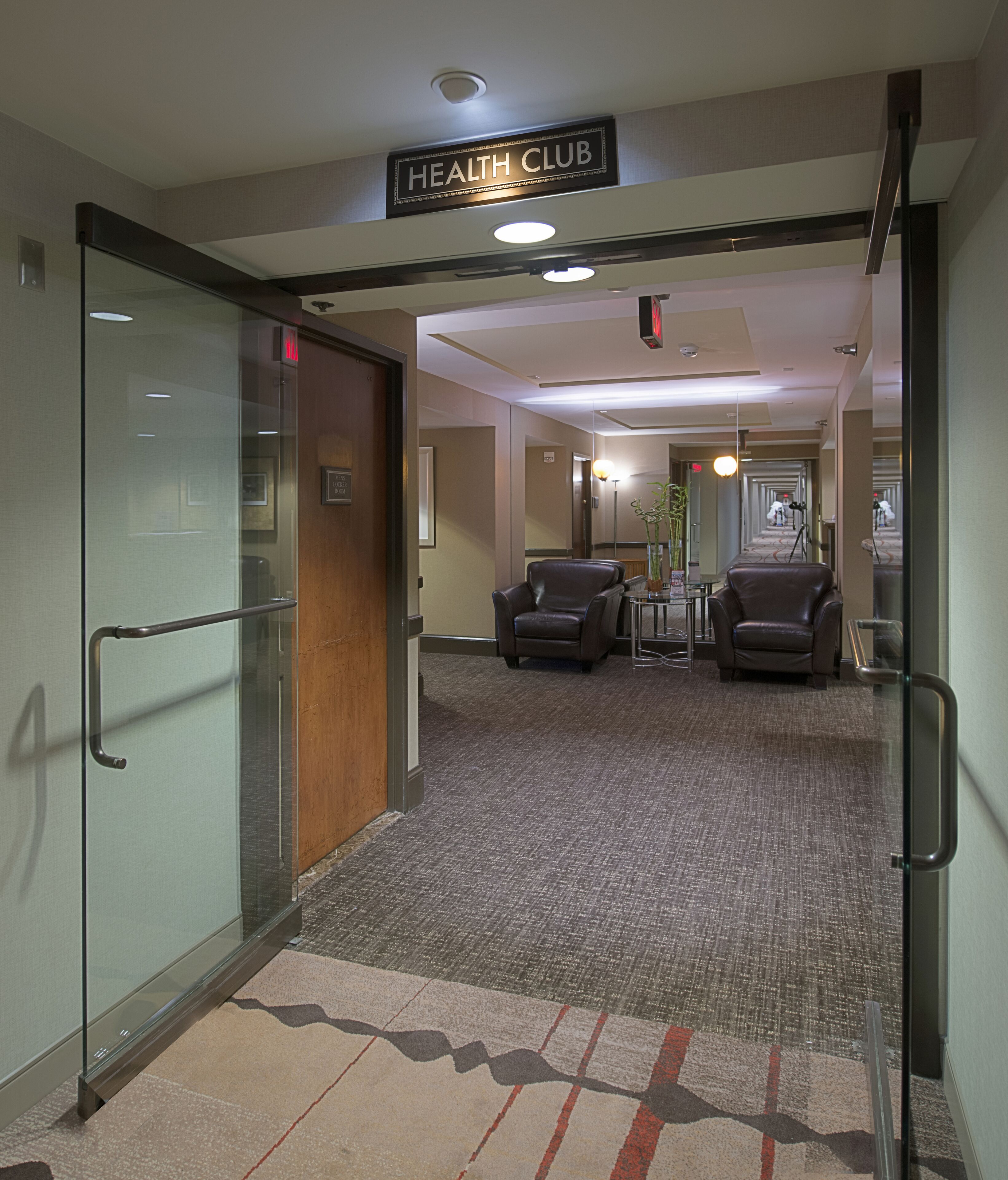 A hotel corridor with a 'HEALTH CLUB' sign, glass doors, and two leather chairs.