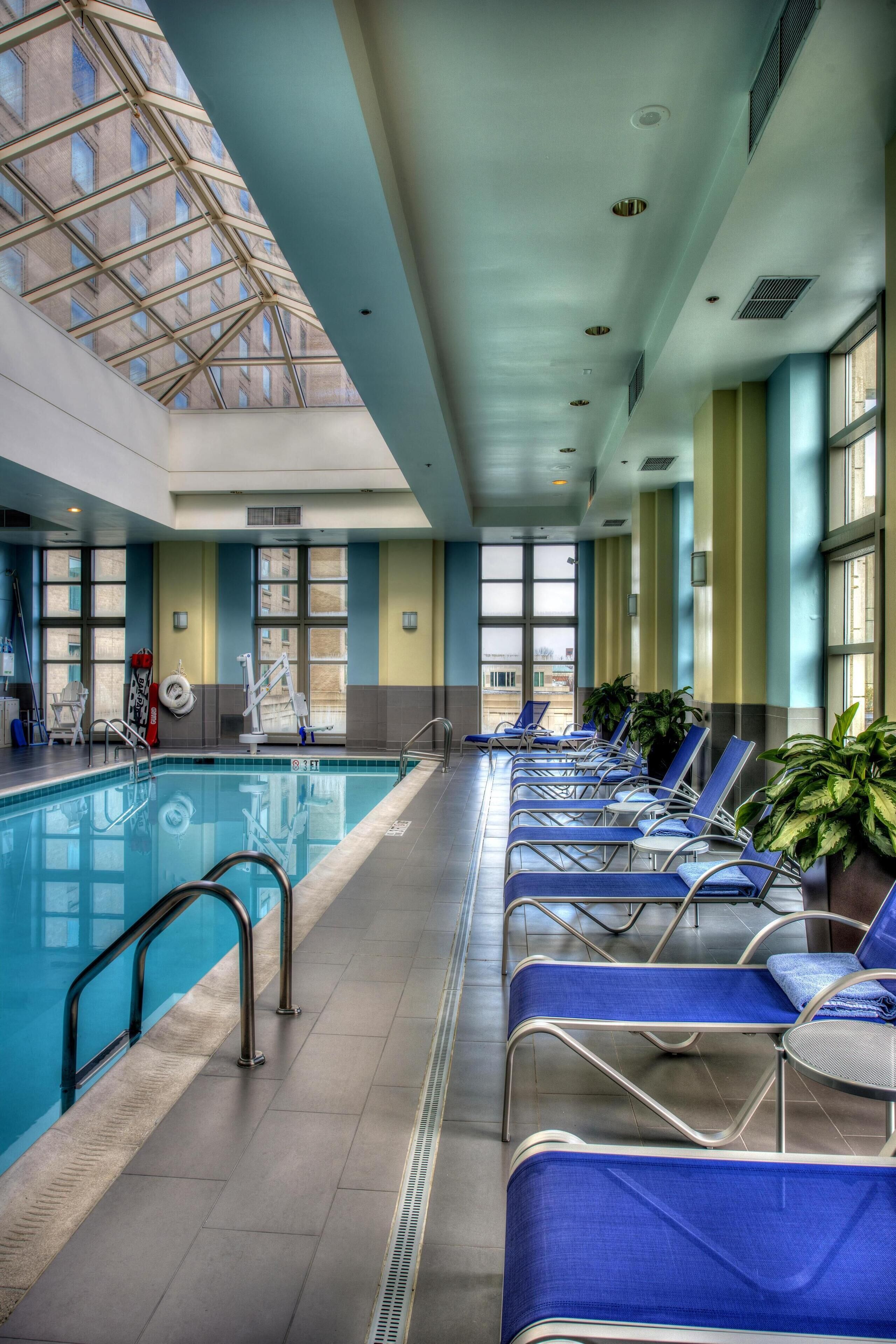An indoor swimming pool with blue lounge chairs, a diving board, and large windows.