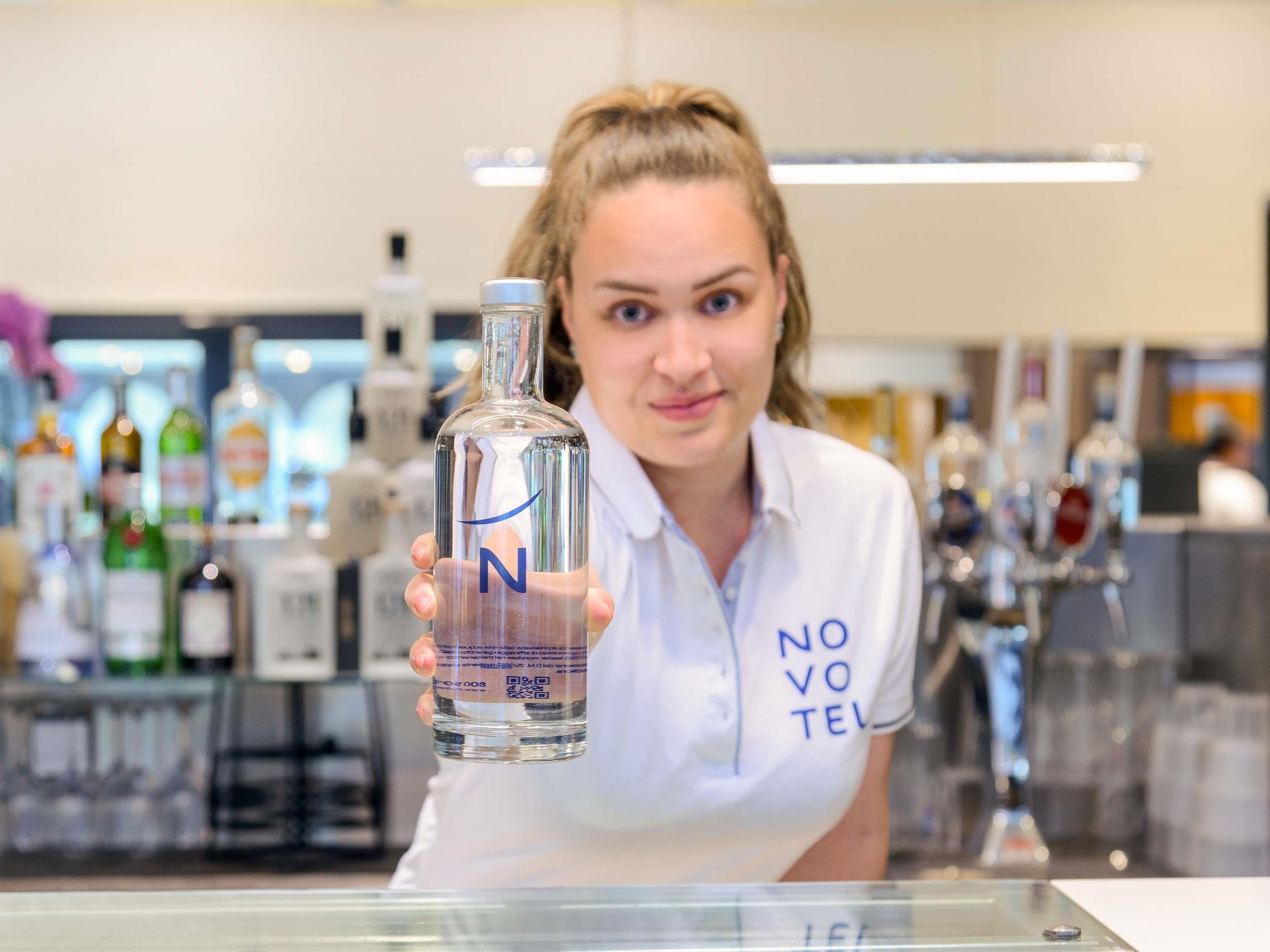 Een persoon houdt een fles met een blauw etiket vast, staand in een bar met verschillende flessen op de achtergrond.