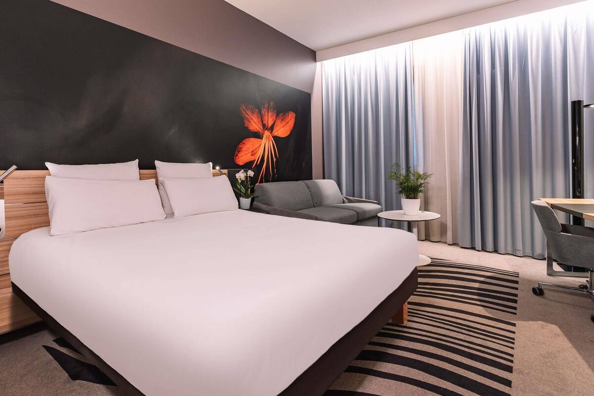 A modern hotel room with a large bed, a grey sofa, a small round table, and a potted plant.
