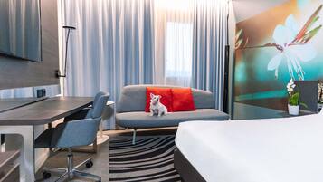 Habitación de hotel con cama, escritorio, silla, sofá con almohada roja, televisor y ventana grande con cortinas.