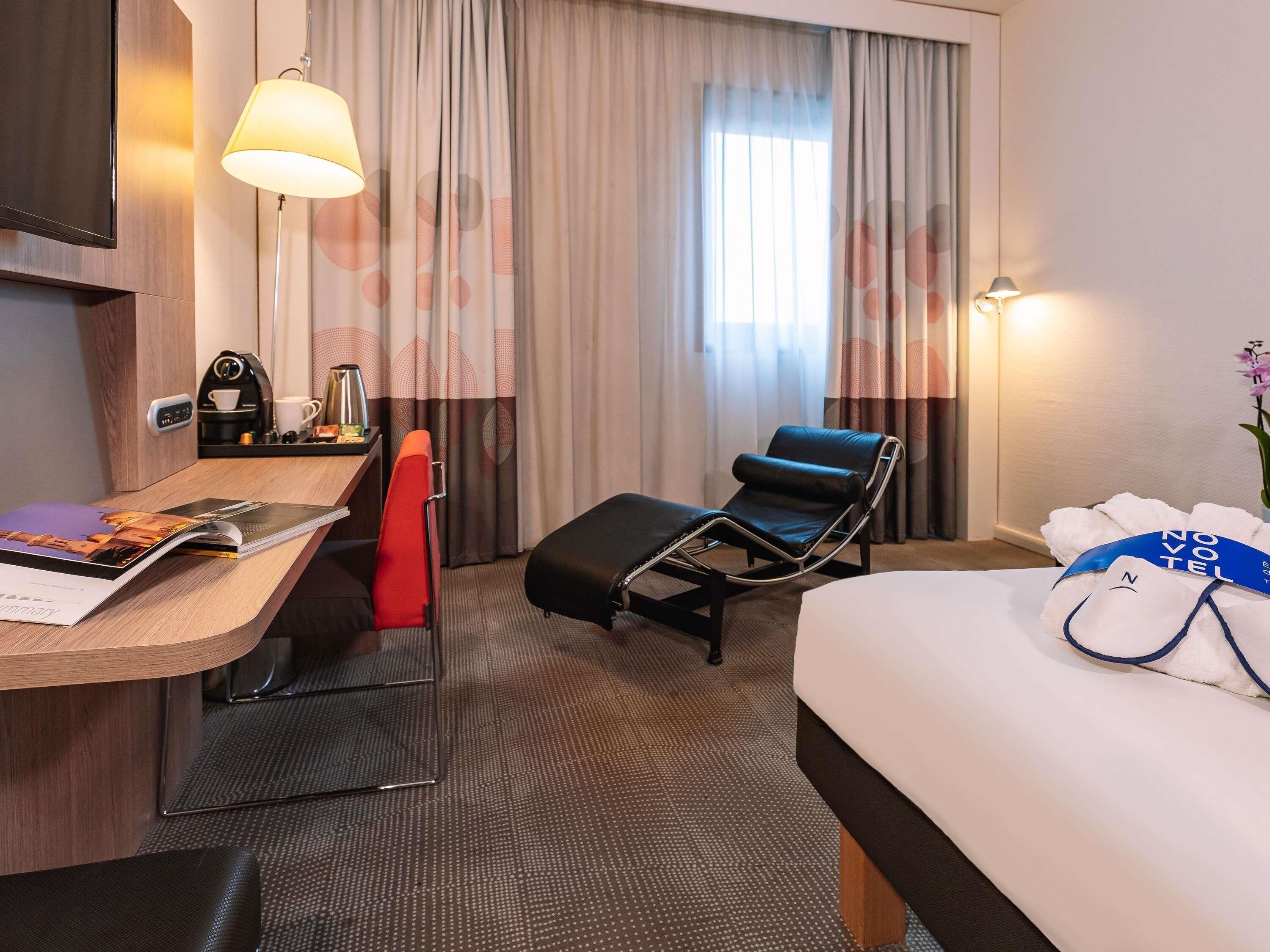 Habitación de hotel con cama, escritorio con lámpara, una silla, cafetera y ventana con cortinas.