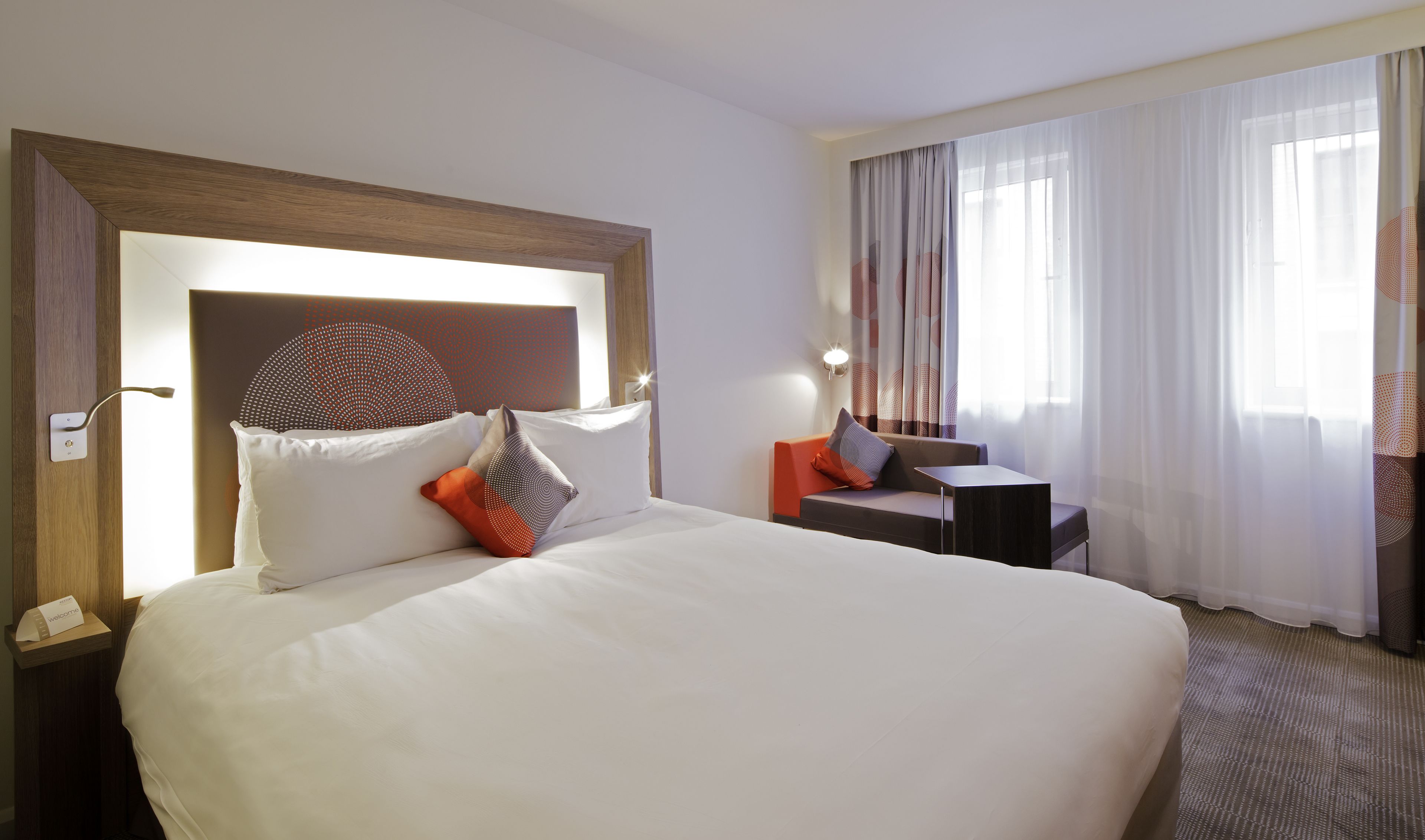 A hotel room with a large bed, a desk, and a view of the outdoors through sheer curtains.