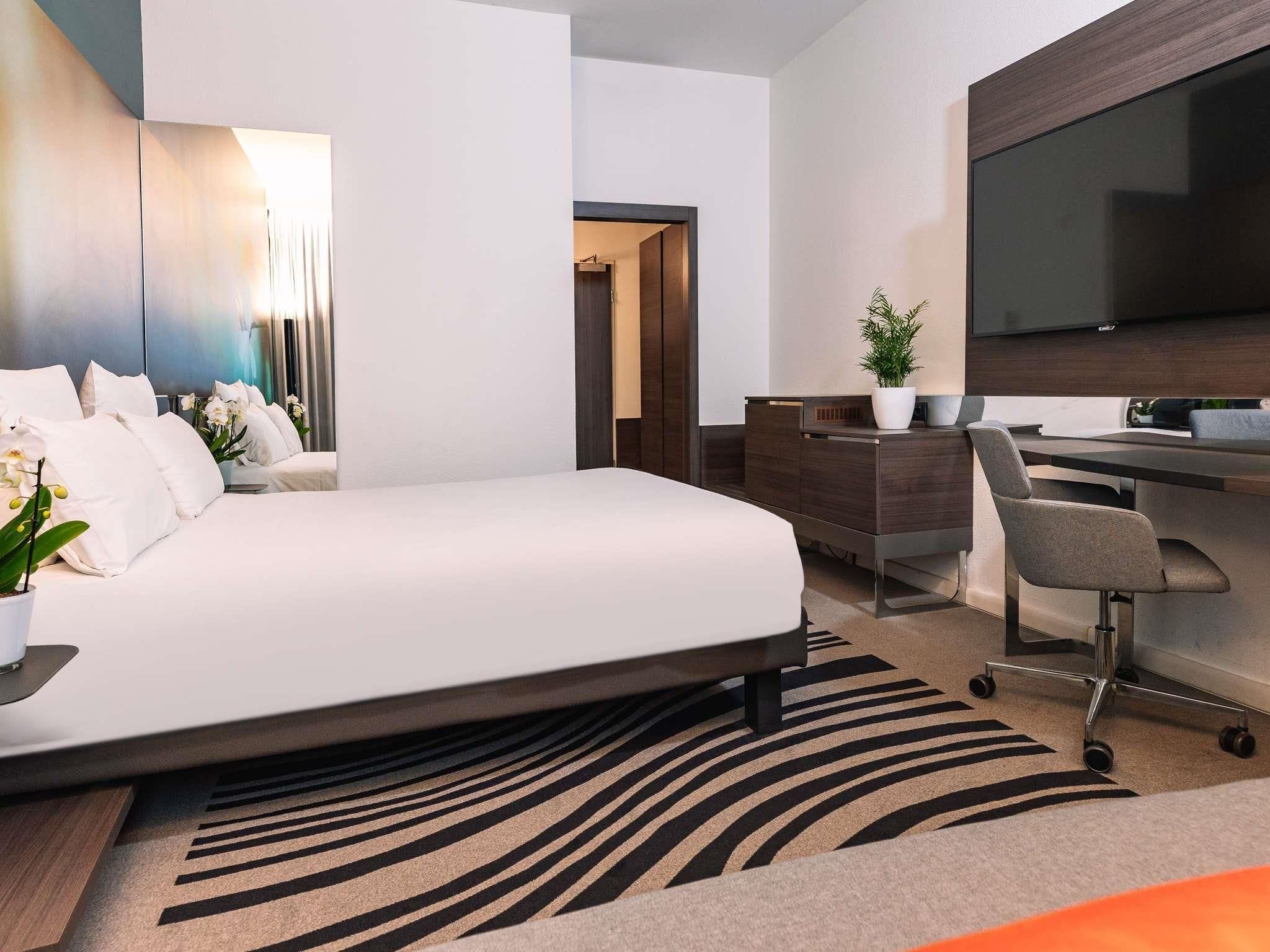 A modern hotel room with a large bed, a desk, a chair, a television, and a potted plant.