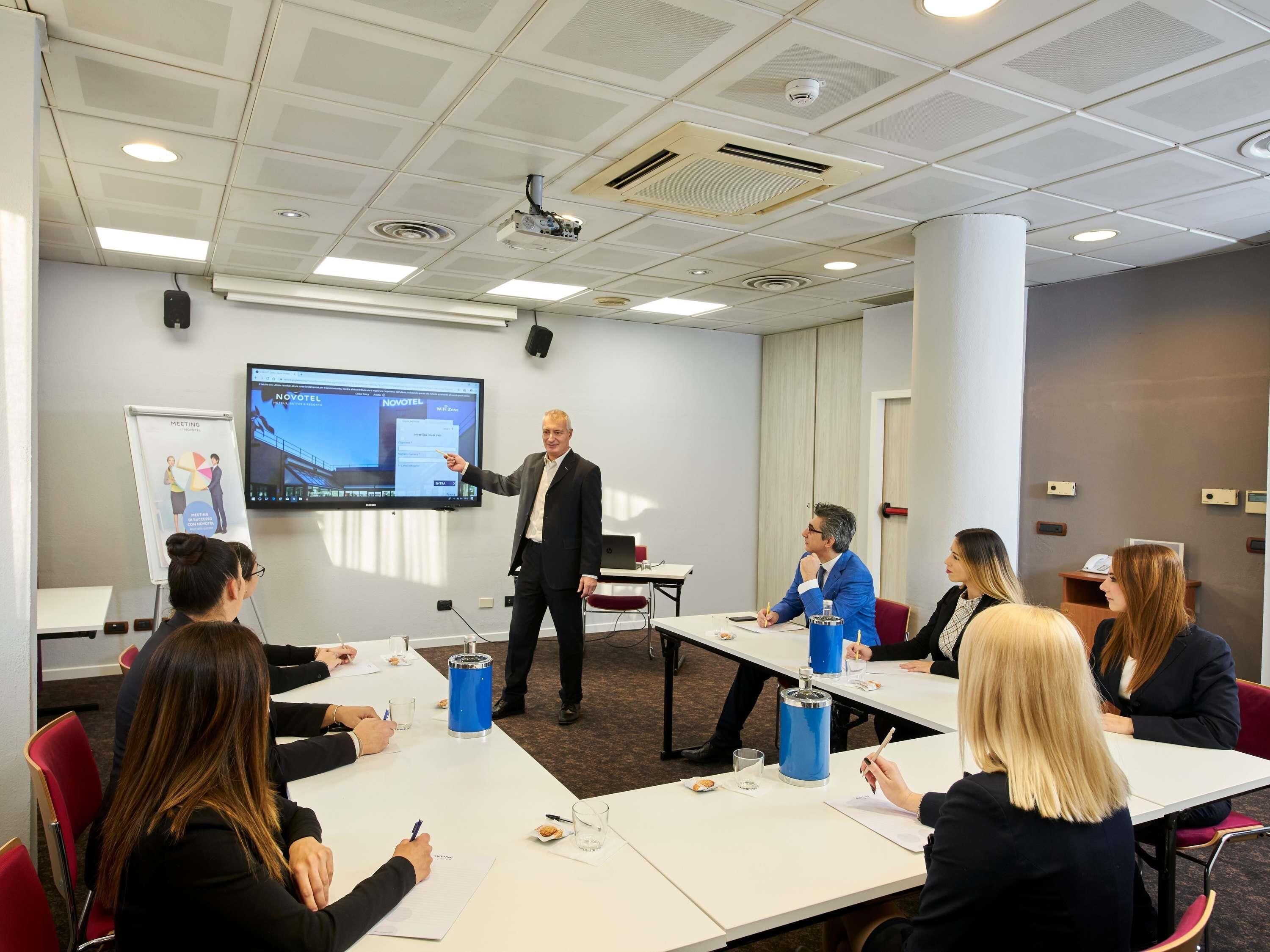 A conference room with a presentation screen, a projector, and attendees seated at tables with notepads and pens.