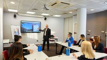 A conference room with a presentation screen, a projector, and attendees seated at tables with notepads and pens.