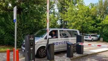 Un veicolo parcheggiato presso una stazione di ricarica, con un segnale 'NOVOTEL', che indica un albergo o una sistemazione nelle vicinanze.