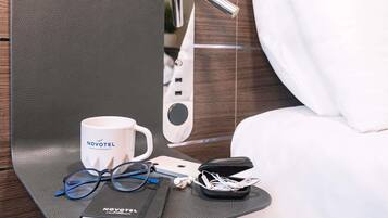 A hotel room with a bed, a bedside table with a white mug, a pair of glasses, a notebook, and a charging cable.