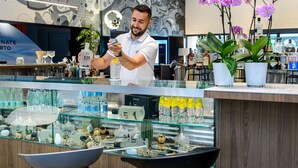 A bar area with a bartender preparing a drink, a display of gin bottles, and decorative items on shelves.